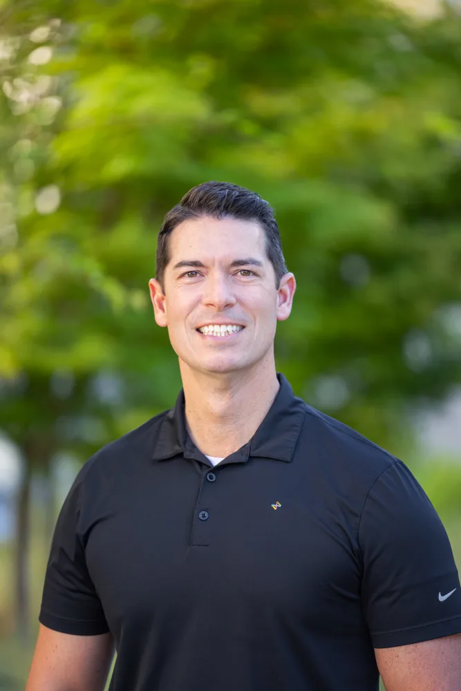 Smiling man in a black polo shirt standing outdoors with green foliage in the background