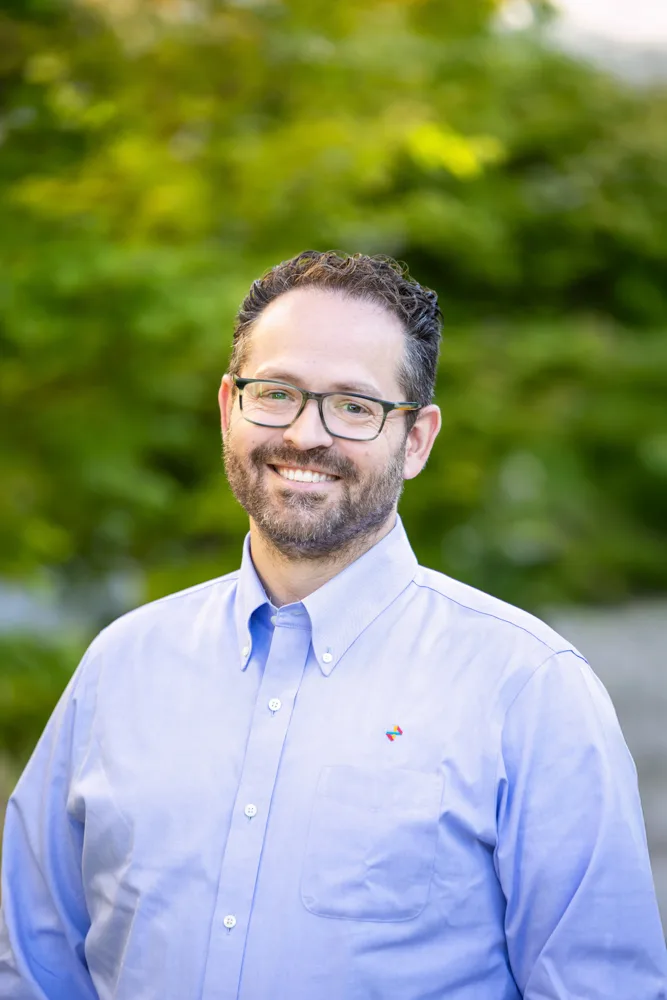 Man with glasses and a short beard smiling in a light blue button-down shirt, standing outdoors with green foliage behind him