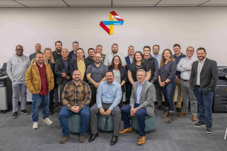 Group of office employees posing together in a conference room beneath a colorful geometric company logo.
