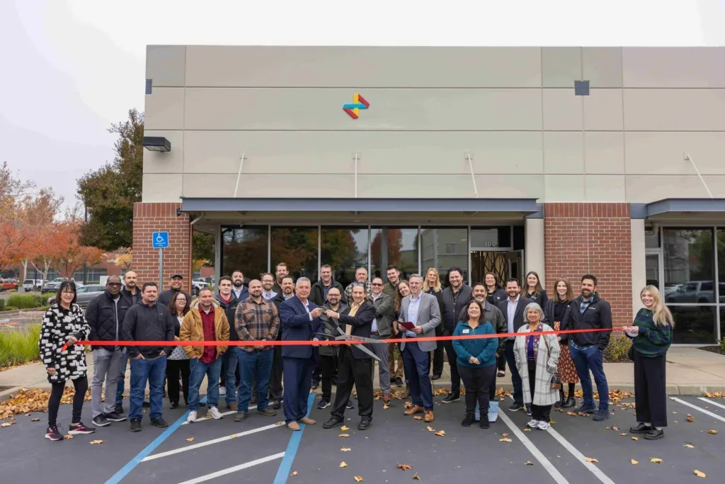 Group of people at a ribbon-cutting ceremony in front of a building, with a red ribbon stretched across the parking lot.