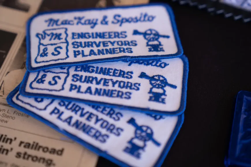 Stack of embroidered “MacKay & Spotto Engineers Surveyors Planners” patches featuring a cartoon surveyor