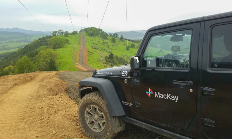 Black Jeep Wrangler with “MacKay” logo parked on a dirt trail overlooking green rolling hills and a powerline road.