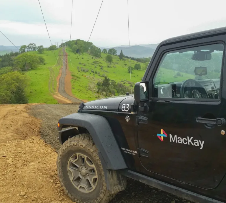 Black Jeep Wrangler with “MacKay” logo parked on a dirt trail overlooking green rolling hills and a powerline road.