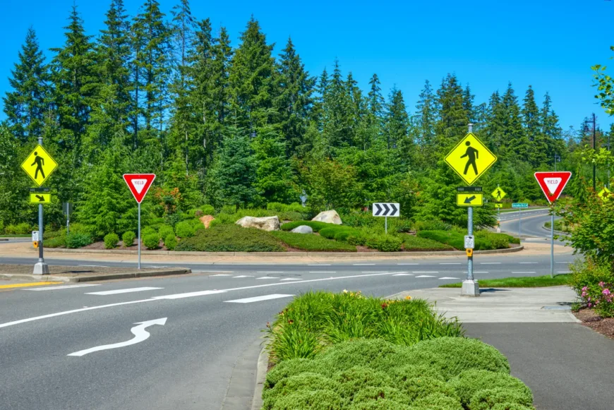Landscaped roundabout with yield signs, pedestrian crossing signs and button, directional arrows on the roadway, and evergreen trees in the background under a clear blue sky.