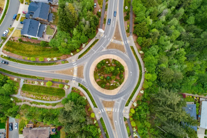 Aerial view of a single-lane three-way roundabout with a landscaped central island, marked crosswalks at each approach, and surrounding suburban homes and dense greenery.