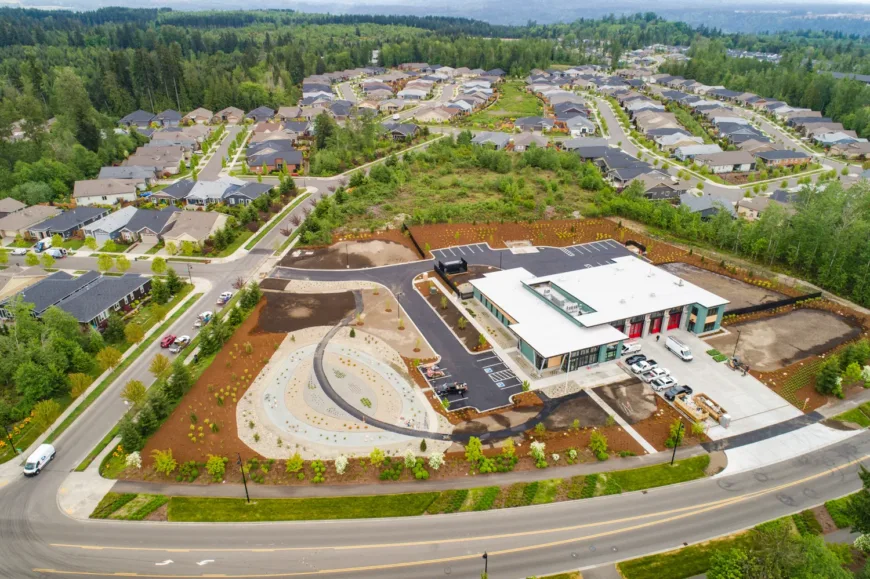 Aerial view of a suburban neighborhood bordered by forest, highlighting a modern single-story building with a white roof, red bay doors, parking lot, and landscaped grounds along curving streets.