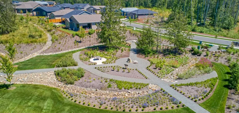 Aerial view of a suburban neighborhood greenbelt with winding gravel walking paths, a circular picnic area, boulders and a dry creek bed amid drought‑tolerant plantings, bordered by lawns and single-story homes near a forest.