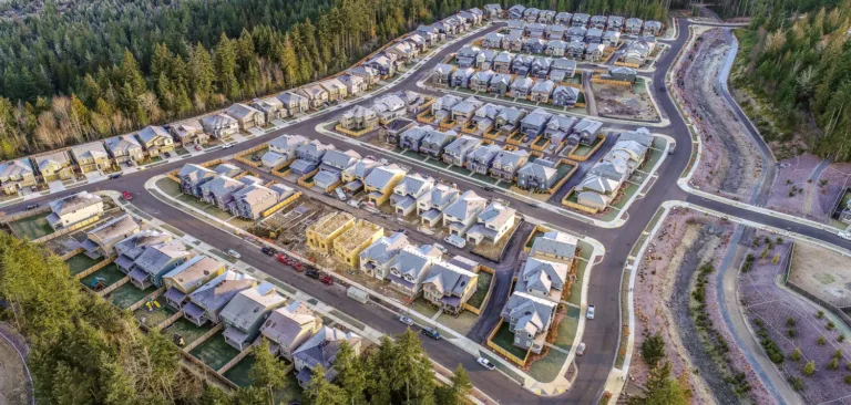 Aerial view of a new suburban neighborhood, with rows of completed and partially framed houses along winding streets, bordered by dense evergreen forest and a drainage corridor.