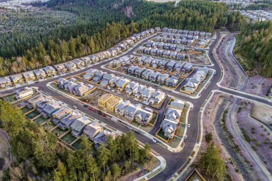 Aerial view of a new suburban neighborhood, with rows of completed and partially framed houses along winding streets, bordered by dense evergreen forest and a drainage corridor.