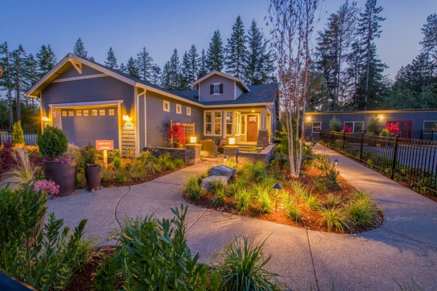 Craftsman-style gray single-story home at dusk with warm exterior lights, a curved concrete walkway through lush landscaped garden beds and potted plants, and tall evergreens in the background.