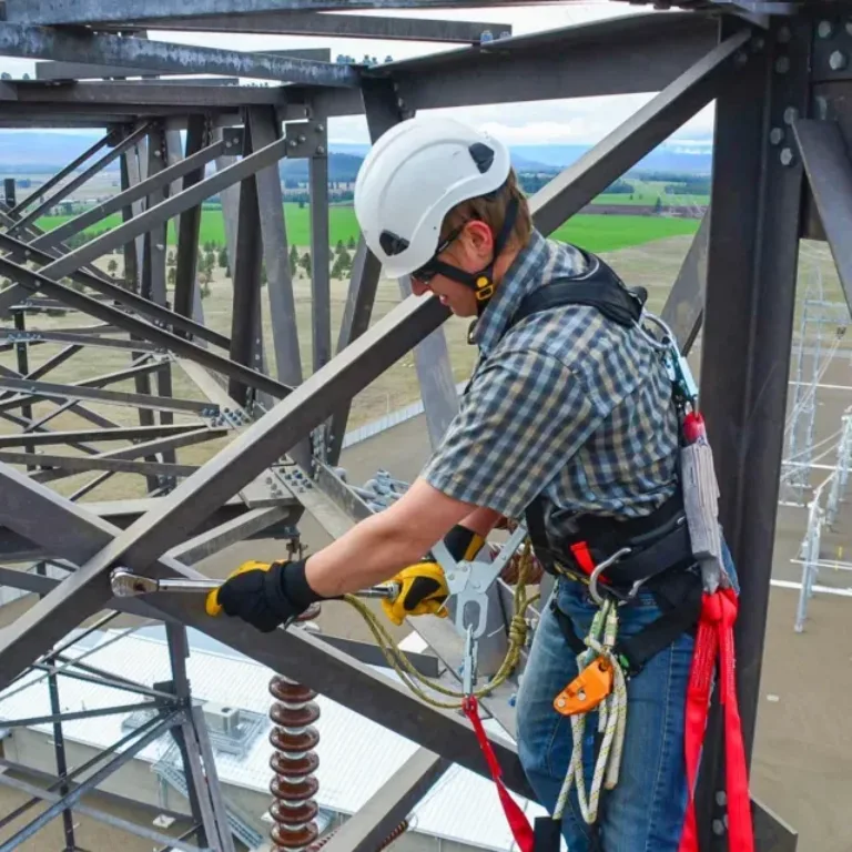 Utility worker in safety gear using a wrench on a high-voltage transmission tower, secured with harness and lanyards above fields.