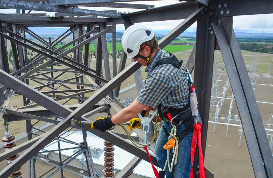 Worker in a safety harness and helmet securing bolts on a high metal transmission tower, with tools clipped and landscape below.