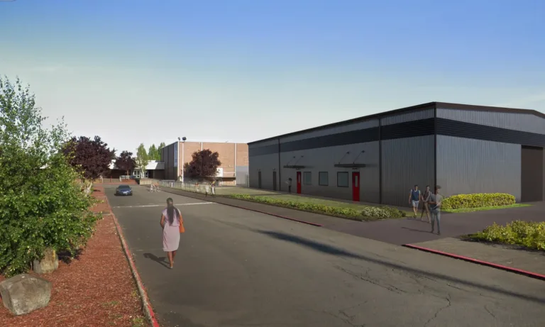 Corrugated metal building with black trim and red doors lines a campus road, with pedestrians, a car, and landscaped beds under a clear blue sky.