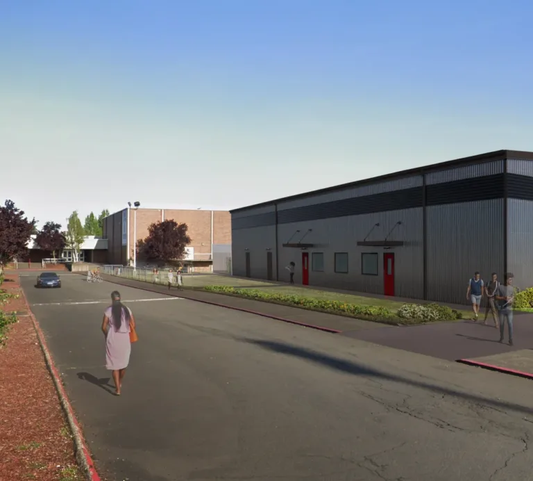 Corrugated metal building with black trim and red doors lines a campus road, with pedestrians, a car, and landscaped beds under a clear blue sky.