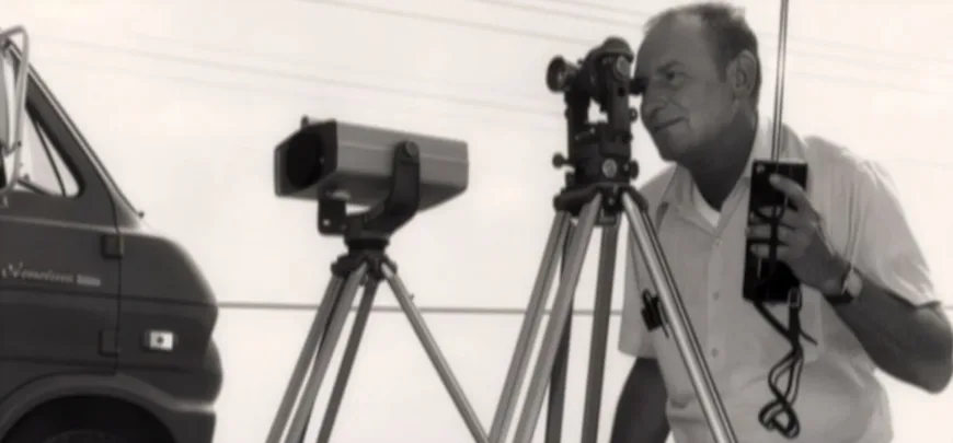 Historical MacKay photo of a surveyor operating tripod-mounted surveying instruments beside a van, in black and white.
