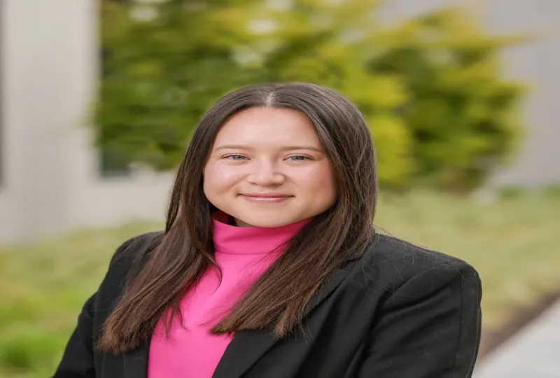 Headshot of Alivia Quitugua, with long straight brown hair, wearing a bright pink turtleneck and black blazer, smiling slightly outdoors with a blurred green background.