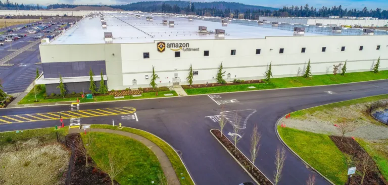 Aerial view of a large Amazon fulfillment center warehouse with a white roof and logo on the facade, surrounded by parking lots, landscaped roads, and a wooded horizon under a cloudy sky.