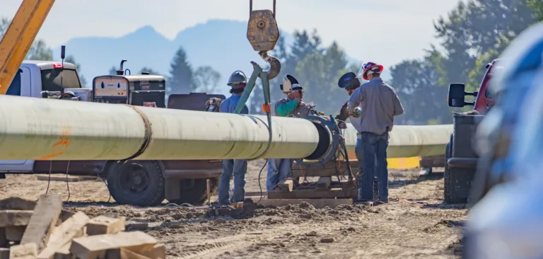 Workers in hard hats weld and align a long green pipeline as a crane suspends a section at a dusty outdoor job site, with trucks, trees, and distant mountains in the background.