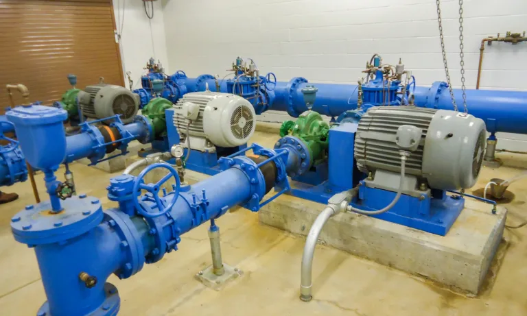 Industrial pump room with multiple blue pipes, valves, and gauges connected to large electric motors driving centrifugal pumps mounted on concrete pads.