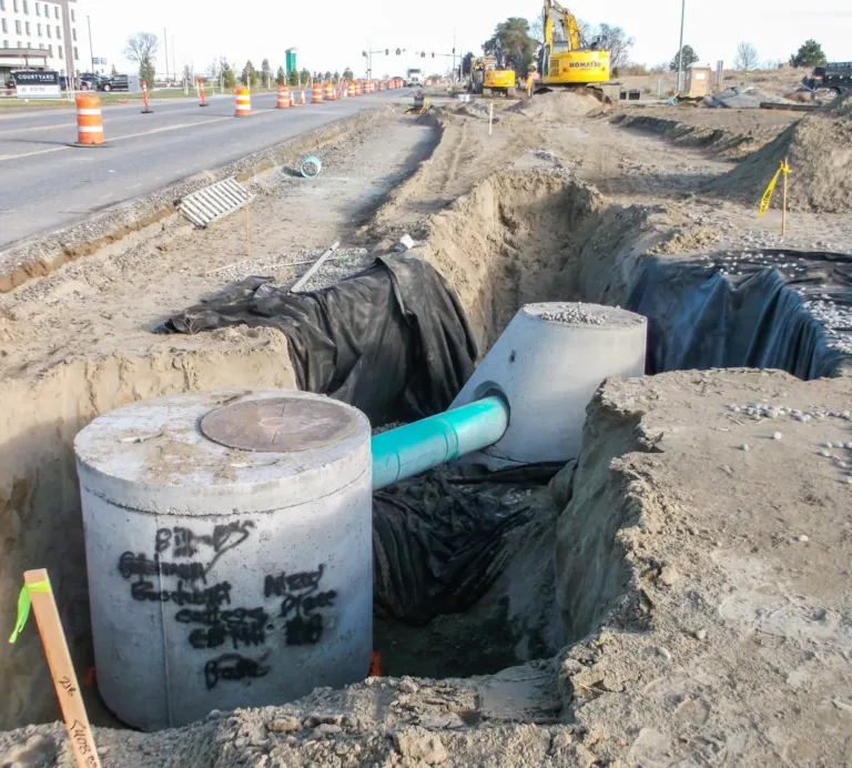 Roadwork scene showing an open trench with concrete storm drain manholes connected by a green pipe beside traffic barrels, excavators, and a city street.