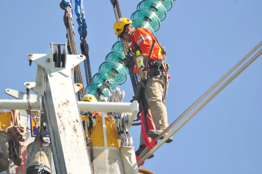 Utility lineworker in helmet and safety harness works on high-voltage transmission hardware beside green glass insulators atop a tower against a clear blue sky.