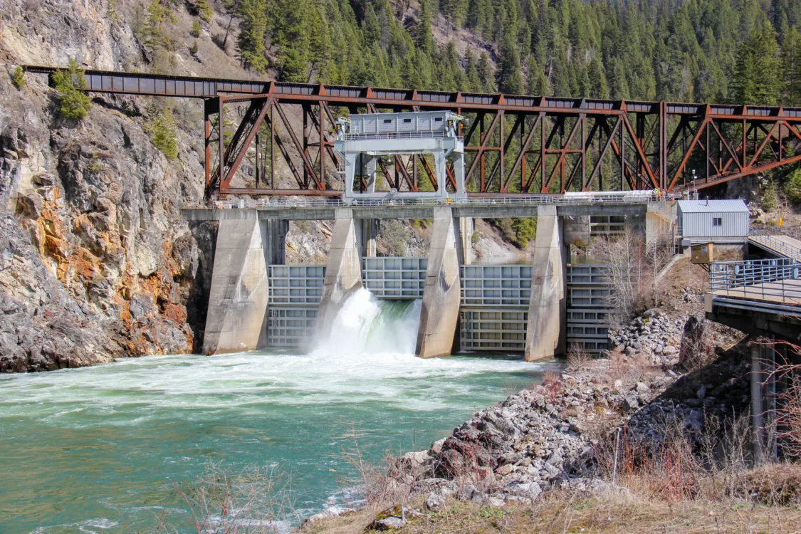 Concrete dam with a spillway releasing water beneath a rusted steel truss railway bridge in a forested mountain gorge.