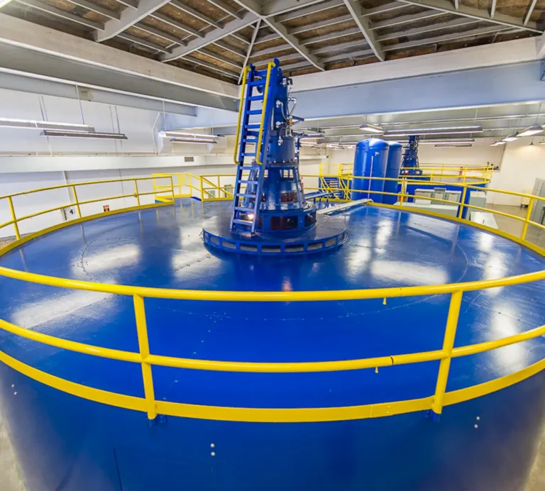 Large blue circular industrial machine with yellow safety railings and a central laddered mechanism inside a brightly lit plant room.