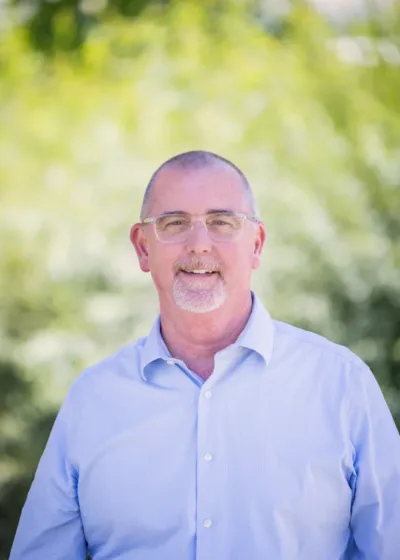 Headshot of Bryan Cole smiling in a light blue button-down shirt, wearing glasses and a short gray goatee, against a blurred green outdoor background.