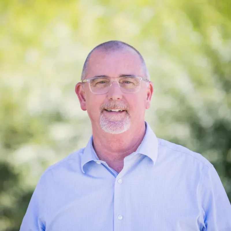 Headshot of Bryan Cole smiling in a light blue button-down shirt, wearing glasses and a short gray goatee, against a blurred green outdoor background.