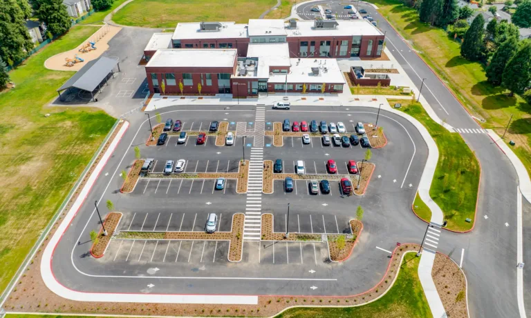 Aerial view of a modern brick school with a large parking lot featuring landscaped islands and crosswalks, a playground and covered area to the left, a looped drop-off drive to the right, and surrounding lawns and trees.