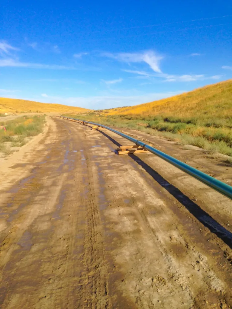 A long green pipeline on wooden supports stretches along a muddy dirt road through rolling grassy hills under a clear blue sky.