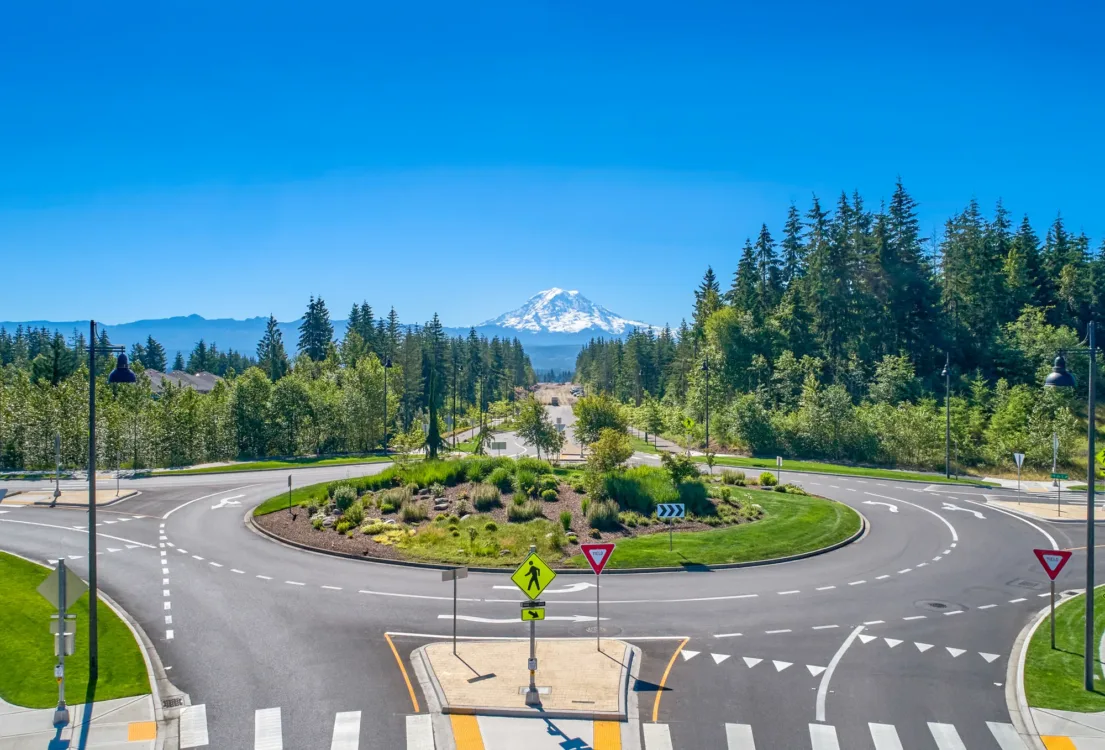 A landscaped roundabout with crosswalks, yield signs, and a pedestrian crossing marker sits amid evergreen trees, with a distant snow-capped mountain under a clear blue sky.