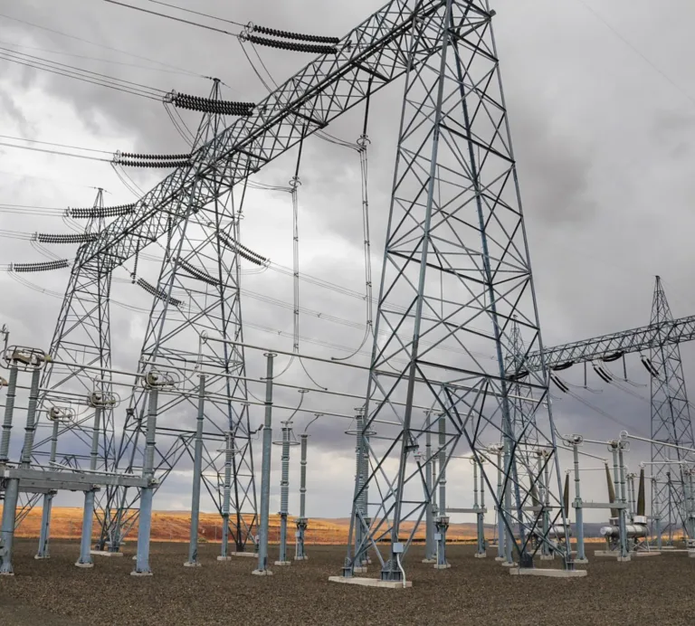 High-voltage electrical substation with lattice towers, insulators, and overhead power lines in an open, arid landscape under cloudy skies.