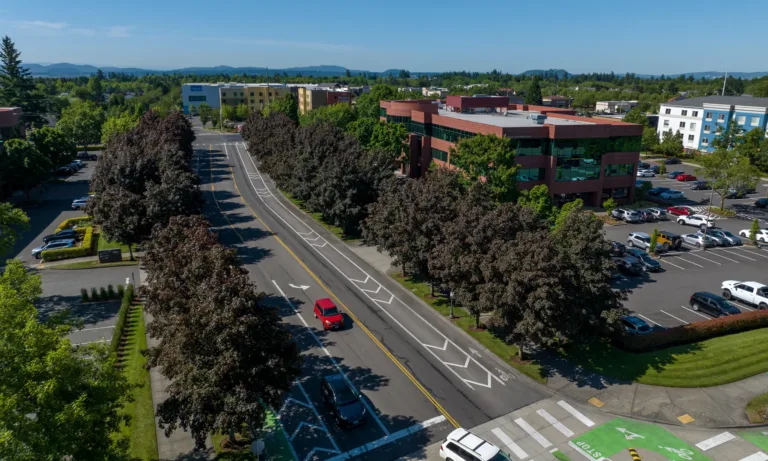 Aerial view of a tree-lined urban street with green-painted bike lanes and a crosswalk, cars traveling past a red brick office building and parking lots under a clear blue sky.