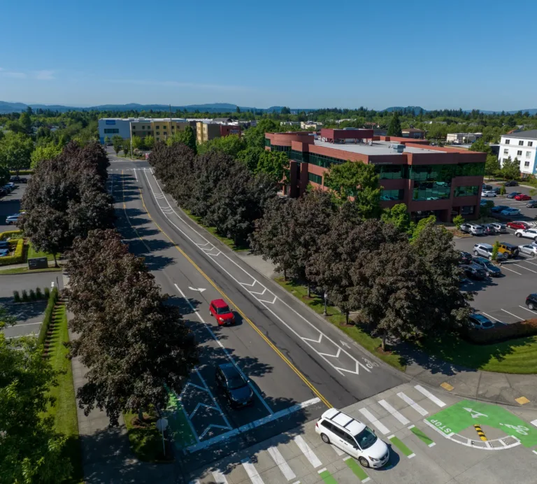 Aerial view of a tree-lined urban street with green-painted bike lanes and a crosswalk, cars traveling past a red brick office building and parking lots under a clear blue sky.