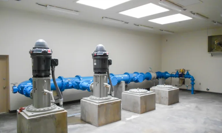 Industrial pump room with three vertical pumps on concrete pedestals connected to a large blue water pipeline beneath bright ceiling panels.