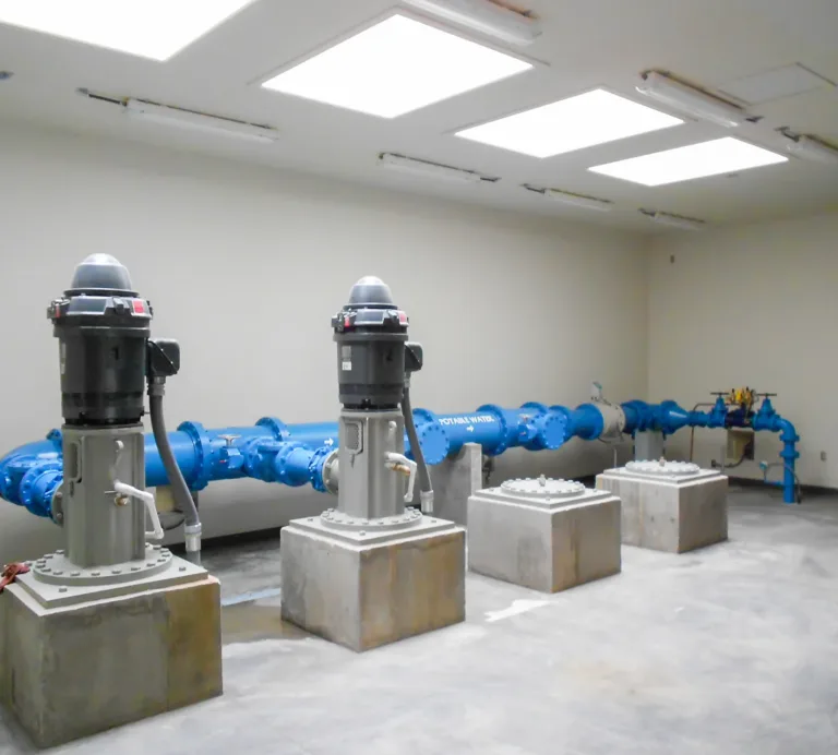 Industrial pump room with three vertical pumps on concrete pedestals connected to a large blue water pipeline beneath bright ceiling panels.