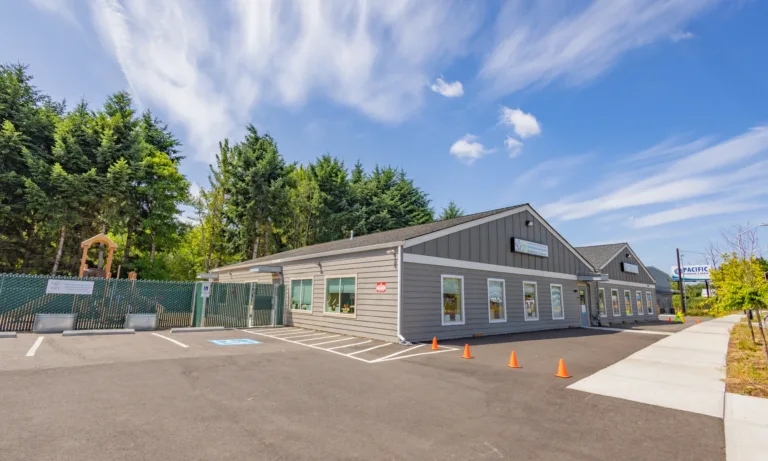 Single-story gray building with multiple gables and large windows, a fenced playground on the left, accessible parking spaces and orange cones in the front lot, bordered by tall evergreens under a bright blue sky.