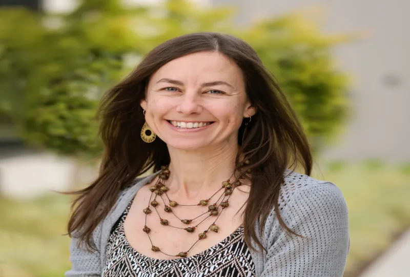 Headshot of Emily Mills smiling with long brown hair, wearing a patterned top, gray knit cardigan, layered beaded necklace, and dangling earrings, outdoors with a soft green background.