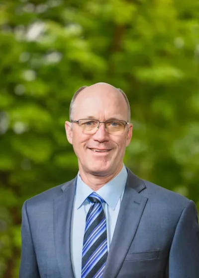 Headshot of Erik Nelson, a smiling man with glasses in a blue suit and striped tie, posed outdoors against a blurred green background.