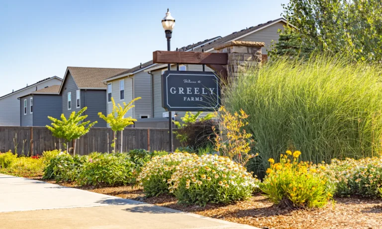 Landscaped neighborhood entrance sign reading “Welcome to Greely Farms” beside ornamental grasses and flowers, with suburban homes and a lamppost under a clear blue sky.