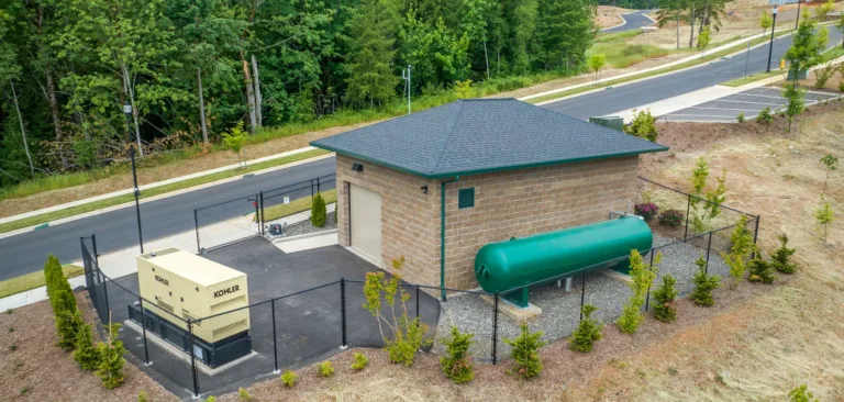 Brick utility building with a chain-link fenced yard housing a large green horizontal fuel tank and a beige generator beside a quiet road and forest.