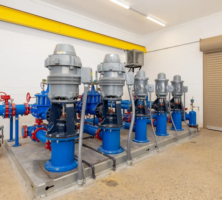 Row of blue industrial pumps with gray vertical motors and red valve wheels mounted on a skid in a clean pump room, with piping, a yellow overhead beam, and a beige roll-up door in the background.