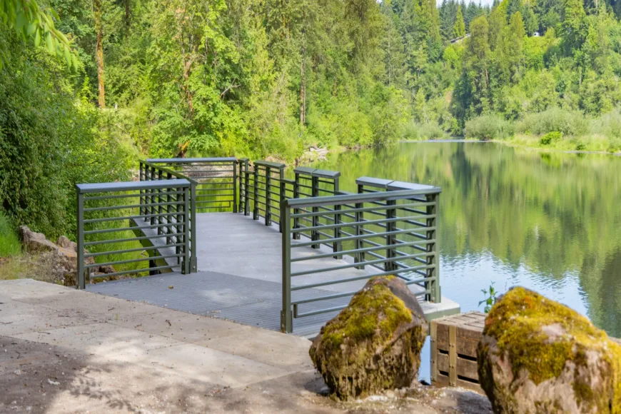 Metal riverside viewing platform with railings extends over a calm, tree-lined river, with mossy boulders in the foreground and forest reflections on the water.