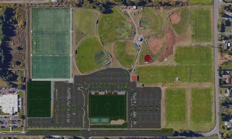 Aerial view of a large sports complex with multiple soccer fields (turf and grass), a cluster of baseball/softball diamonds, extensive central parking lots, and surrounding roads and trees.