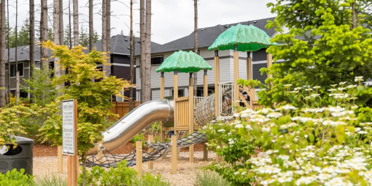 Community playground with green canopy-topped structures, a metal tube slide, and rope bridges, surrounded by landscaped greenery with townhomes and tall trees in the background.