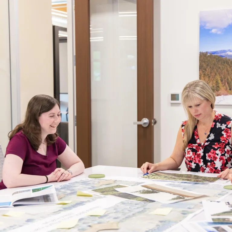 Three colleagues in a modern conference room review maps and documents covered with sticky notes, discussing plans around a white table beneath a wall photo of a snowy mountain landscape.