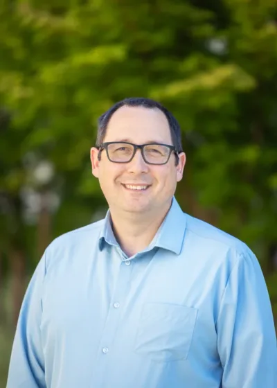 Headshot of Jerry Balch, a smiling person with short dark hair and glasses wearing a light blue button-down shirt against a blurred green foliage background.