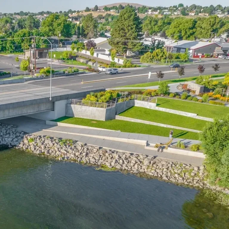 Riverside park with walking paths and landscaped lawns beneath a road bridge, with a neighborhood and hills in the background