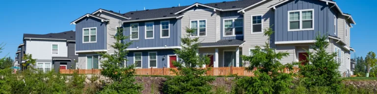 Row of modern two-story townhomes with gray and blue siding, red doors, and gabled roofs, set behind a wooden fence with young evergreens and rock landscaping under a clear blue sky.